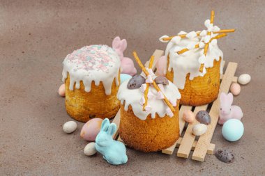 Traditional Easter cakes with white frosting and decorative pussy willow branches on wooden stand. Festive still life with pastel eggs and bunny figures on stone background, copy space