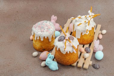 Traditional Easter cakes with white frosting and decorative pussy willow branches on wooden stand. Festive still life with pastel eggs and bunny figures on stone background, copy space