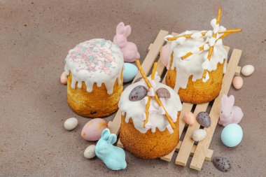 Traditional Easter cakes with white frosting and decorative pussy willow branches on wooden stand. Festive still life with pastel eggs and bunny figures on stone background, copy space