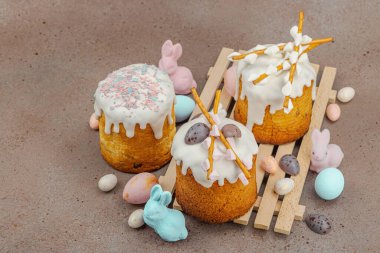 Traditional Easter cakes with white frosting and decorative pussy willow branches on wooden stand. Festive still life with pastel eggs and bunny figures on stone background, copy space