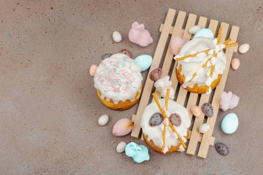 Traditional Easter cakes with white frosting and decorative pussy willow branches on wooden stand. Festive still life with pastel eggs and bunny figures on stone background, top view
