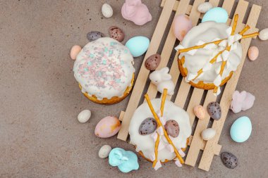 Traditional Easter cakes with white frosting and decorative pussy willow branches on wooden stand. Festive still life with pastel eggs and bunny figures on stone background, top view