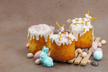 Traditional Easter cakes with white frosting and decorative pussy willow branches on wooden stand. Festive still life with pastel eggs and bunny figures on stone background, copy space