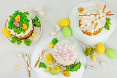 Beyaz kremalı Paskalya pastası, yosun süslemesi ve beyaz tabakta bıldırcın yumurtası. Renkli tavşancıklar ve açık gri arka planda yenilebilir söğüt dalları ile neşeli bahar kompozisyonu, üst görünüm