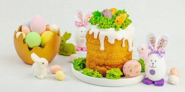 Beyaz kremalı Paskalya pastası, yosun süslemesi ve beyaz tabakta bıldırcın yumurtası. Renkli tavşancıklar ve açık gri arka planda yenilebilir söğüt dalları ve pankart formatlı şenlikli bahar kompozisyonu