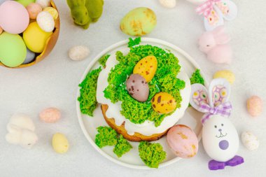 Beyaz kremalı Paskalya pastası, yosun süslemesi ve beyaz tabakta bıldırcın yumurtası. Renkli tavşancıklar ve açık gri arka planda yenilebilir söğüt dalları ile neşeli bahar kompozisyonu, üst görünüm