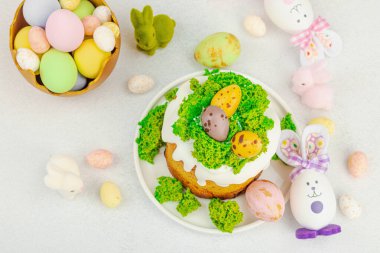 Beyaz kremalı Paskalya pastası, yosun süslemesi ve beyaz tabakta bıldırcın yumurtası. Renkli tavşancıklar ve açık gri arka planda yenilebilir söğüt dalları ile neşeli bahar kompozisyonu, üst görünüm
