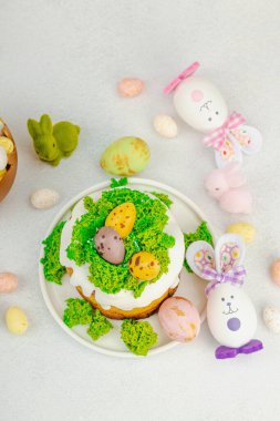 Beyaz kremalı Paskalya pastası, yosun süslemesi ve beyaz tabakta bıldırcın yumurtası. Renkli tavşancıklar ve açık gri arka planda yenilebilir söğüt dalları ile neşeli bahar kompozisyonu, üst görünüm