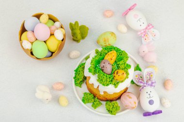 Beyaz kremalı Paskalya pastası, yosun süslemesi ve beyaz tabakta bıldırcın yumurtası. Renkli tavşancıklar ve açık gri arka planda yenilebilir söğüt dalları ile neşeli bahar kompozisyonu, üst görünüm