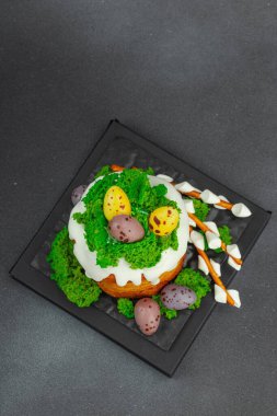 Yenilebilir yosun yuvası, çikolata yumurtası ve el yapımı söğüt dallarıyla süslenmiş Paskalya pastası. Siyah taş arka planda tavşan figürlü geleneksel şenlikli tatlı.