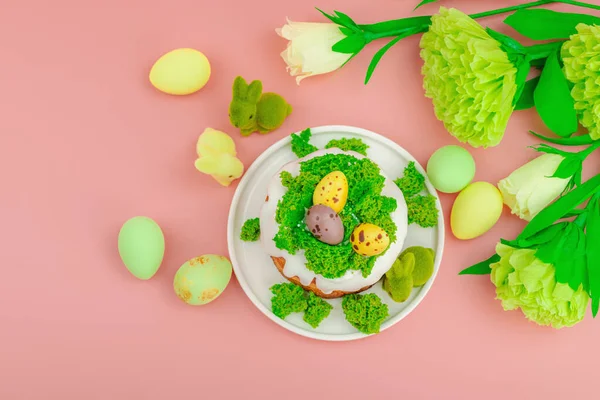 Geleneksel Paskalya pastaları, beyaz kremalı ve tabakta renkli yumurtalar serpiştirilmiş. Pembe arka planda tavşanlı ve çiçekli şenlikli kompozisyon. Ev yapımı tatil pişirme konsepti, üst görünüm