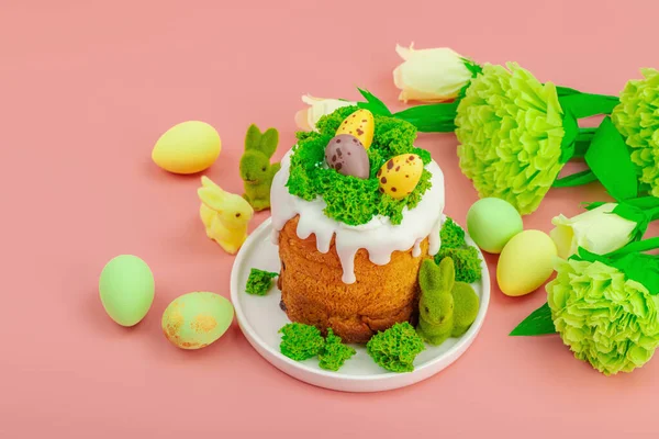 Geleneksel Paskalya pastaları, beyaz kremalı ve tabakta renkli yumurtalar serpiştirilmiş. Pembe arka planda tavşanlı ve çiçekli şenlikli kompozisyon. Ev yapımı tatil pişirme konsepti, kopyalama alanı