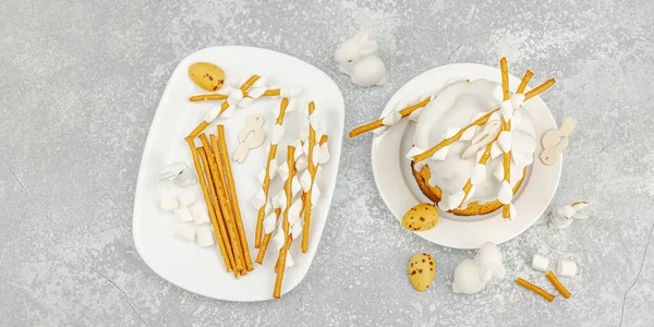 Paskalya pastası dekorasyonu için ekmek çubukları ve lokum kullanarak yenilebilir söğüt dalları yapma süreci. Tatil malzemeleri, bıldırcın yumurtaları ve taş arka planda tavşan figürleri, afiş formatı