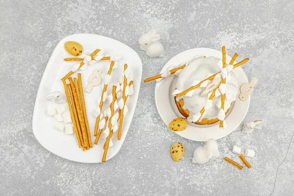 Paskalya pastası dekorasyonu için ekmek çubukları ve lokum kullanarak yenilebilir söğüt dalları yapma süreci. Tatil malzemeleri, bıldırcın yumurtaları ve taş arka planda tavşan figürleri.