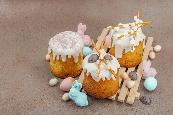 Traditional Easter cakes with white frosting and decorative pussy willow branches on wooden stand. Festive still life with pastel eggs and bunny figures on stone background, copy space