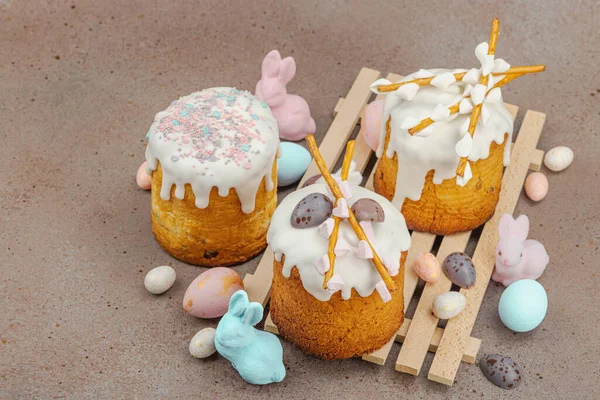 Traditional Easter cakes with white frosting and decorative pussy willow branches on wooden stand. Festive still life with pastel eggs and bunny figures on stone background, copy space