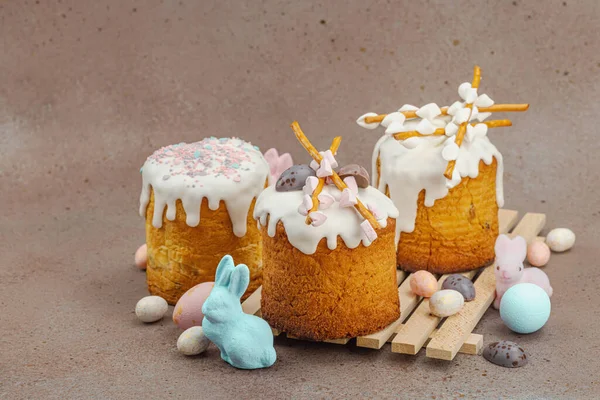 Traditional Easter cakes with white frosting and decorative pussy willow branches on wooden stand. Festive still life with pastel eggs and bunny figures on stone background, copy space
