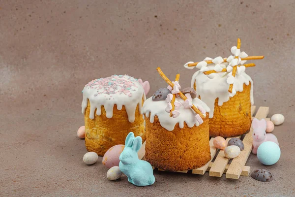 Traditional Easter cakes with white frosting and decorative pussy willow branches on wooden stand. Festive still life with pastel eggs and bunny figures on stone background, copy space