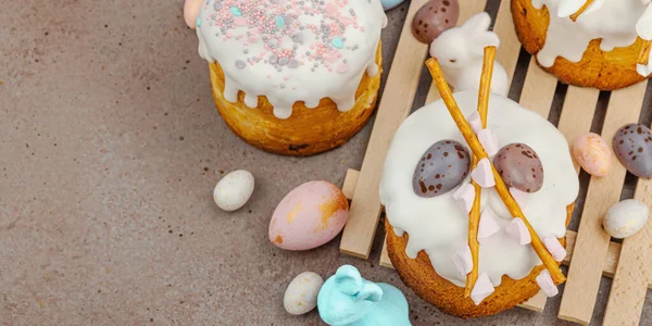 Traditional Easter cakes with white frosting and decorative pussy willow branches on wooden stand. Festive still life with pastel eggs and bunny figures on stone background, banner format