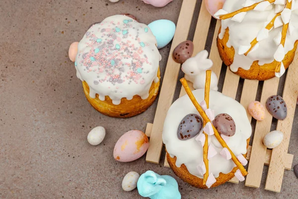 Traditional Easter cakes with white frosting and decorative pussy willow branches on wooden stand. Festive still life with pastel eggs and bunny figures on stone background, top view