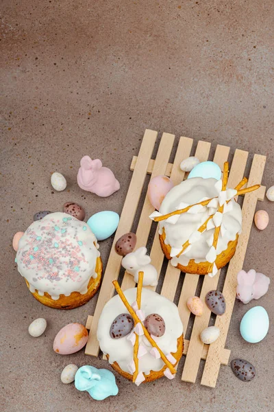 Traditional Easter cakes with white frosting and decorative pussy willow branches on wooden stand. Festive still life with pastel eggs and bunny figures on stone background, top view