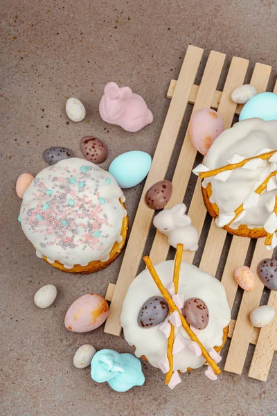 Traditional Easter cakes with white frosting and decorative pussy willow branches on wooden stand. Festive still life with pastel eggs and bunny figures on stone background, top view