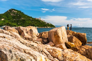 İnsanlar Hong Kong 'daki Stanley Körfezi kıyısındaki kayalıklarda dinleniyor. Su, dağlar, kayalar ve binalarla güzel manzaralı günbatımı manzarası.