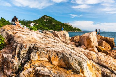 İnsanlar Hong Kong 'daki Stanley Körfezi kıyısındaki kayalıklarda dinleniyor. Su, dağlar, kayalar ve binalarla güzel manzaralı günbatımı manzarası