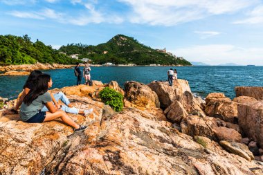 İnsanlar Hong Kong 'daki Stanley Körfezi kıyısındaki kayalıklarda dinleniyor. Su, dağlar, kayalar ve binalarla güzel manzaralı günbatımı manzarası.