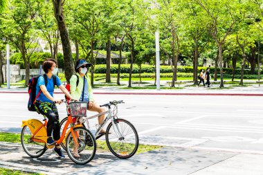 Maske takan insanlar Taipei 101 gökdeleninin önünden bisikletle geçiyorlar. Coronavirus (COVID-19) salgını sırasında.