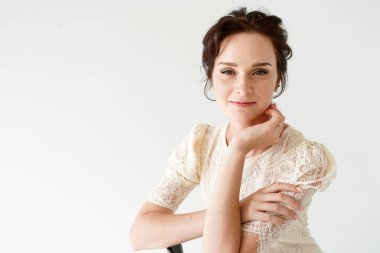 Güzel genç bayan beyaz stüdyoda oturuyor ve kameraya bakıyor, nazikçe gülümsüyor.