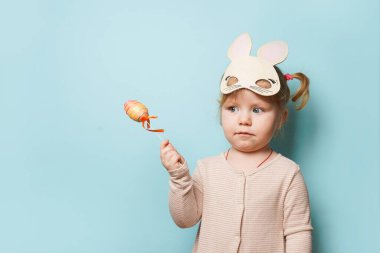 Paskalya tavşanı kulakları takmış şirin bir kızın portresi. Mavi arka planda renkli bir yumurta tutuyor.