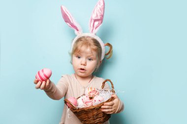 Paskalya tavşanı kulakları takmış, mavi arka planda renkli yumurtalar tutan küçük şirin bir kızın portresi.