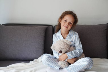 A little girl in blue and white striped pajamas sits on a bed holding a fluffy teddy bear and smiling softly