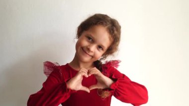 A smiling little girl in a red dress forms a heart shape with her hands, expressing love and happiness