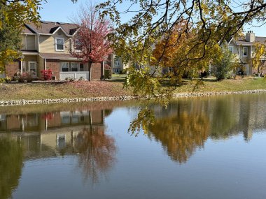 Durgun bir sonbahar sahnesi. Etrafı renkli ağaçlarla çevrili sakin bir gölette yansıyan evler.