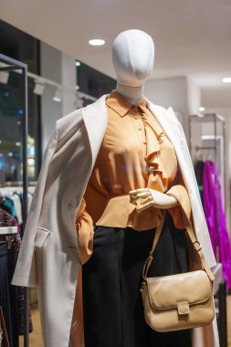 Markette bej bluz, beyaz önlük ve çanta giymiş zarif bir kadın manken.