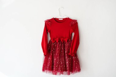 Kırmızı, uzun kollu bir çocuk elbisesi beyaz bir duvara asılı. Tüylü eteği ve dekoratif yayları var.