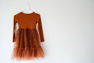 Pas rengi uzun kollu bir çocuk elbisesi beyaz bir duvarda asılı, küçük yıldızlarla süslenmiş parlak tüllü bir etek var.