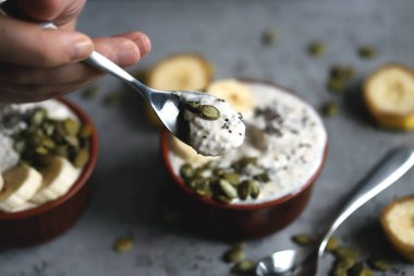 Erkek eli bir kase yoğurt tutuyor. Kaşıklı bir adam tohumlu yoğurt yer. Kabak çekirdeği ve muzlu Chia pudingi. Sağlıklı kahvaltı ya da atıştırmalık. Keto tatlı..