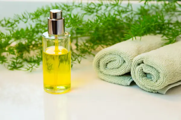 Clear glass spray bottle with yellow liquid. Rolled green towels in a spa setting. Green plant decor in background. Bathroom white countertop.