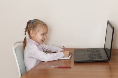 Elinde kalem olan küçük sarışın kız dikkatle dizüstü bilgisayar ekranına bakıyor. Çocuk bilgisayarın önünde uzaktan öğreniyor.