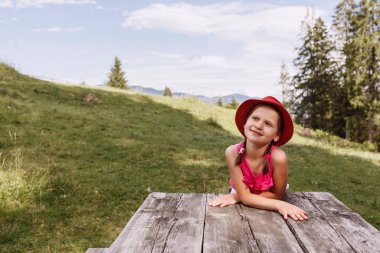 Çimenlikteki ahşap masada oturan kırmızı şapkalı küçük kız. Çocuk dağlarda seyahat eder. Çayırların ve gökyüzünün arka planında pembeli bir bebek