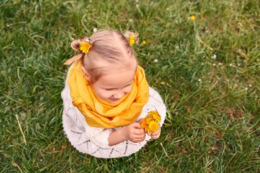 Sarı eşarplı, ellerinde ve saçlarında karahindiba çiçekleri olan küçük bir kız. Çimenlerin üzerinde çiçekli bir çocuk