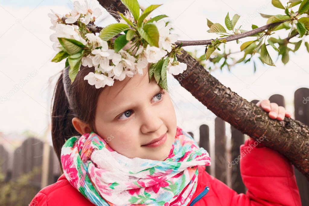 A little girl with a bright scarf looks dreamily into the distance. Child near a flowering cherry branch.