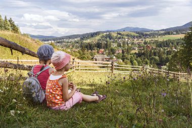 İki çocuk bir dağ açıklığında oturuyor. Tişörtlü iki küçük kız çimlerin üzerinde oturuyor ve dağlara bakıyor. Çiçeklerin arasındaki çimenlikte çocuklar..
