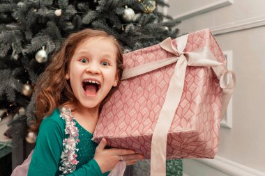 The child is happy with the gift. Happy girl with a gift in her hands on the background of the Christmas tree. Gift box in the hands of a little joyful girl clipart