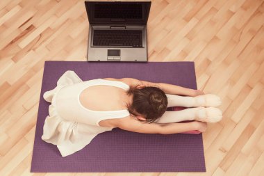 Evde dizüstü bilgisayarın önünde antrenman yapan küçük bir kız. Küçük balerin yerde geriniyor.