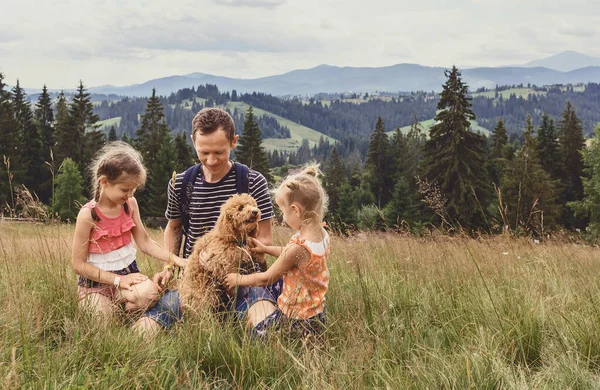 Dad with two little daughters and a dog on a glade on a background of mountains. Family with a little dog on a hike