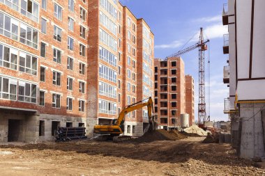 Gökyüzünün arka planında, gökyüzü bulutlu bir binanın yakınındaki kule vinci ve kazıcı. Yüksek tuğlalı bir binanın inşaat alanı
